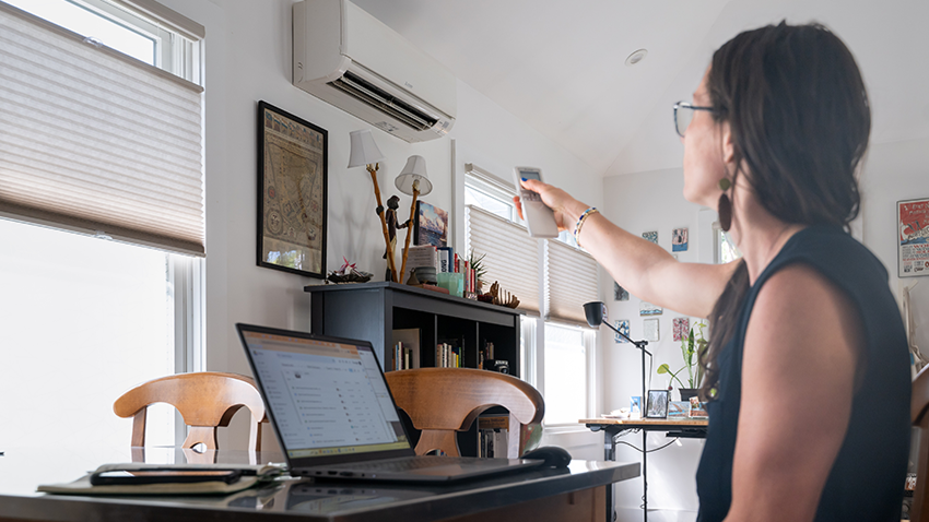Person uses a remote control to operate a wall-mounted heat pump in a home office.