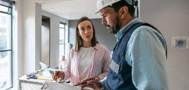 Person in a hard hat reviews paperwork on a clipboard while speaking with a homeowner during an indoor project consultation.