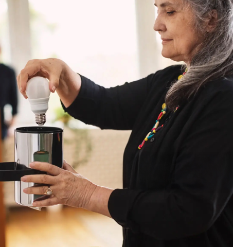 A woman with gray hair installing an energy-efficient LED light bulb into a silver table lamp.