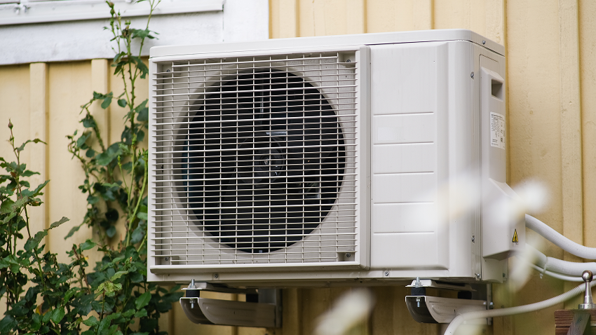 Outdoor heat pump condenser unit mounted on the exterior wall of a building, surrounded by plants.